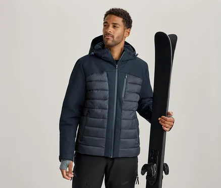 Homme portant une veste de ski bleu foncé et grise et un pantalon de ski noir, tenant une paire de skis noirs.
