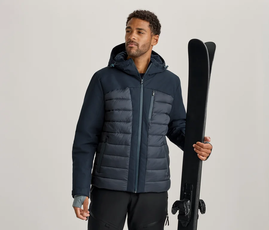 Homme portant une veste de ski bleu foncé et grise et un pantalon de ski noir, tenant une paire de skis noirs.