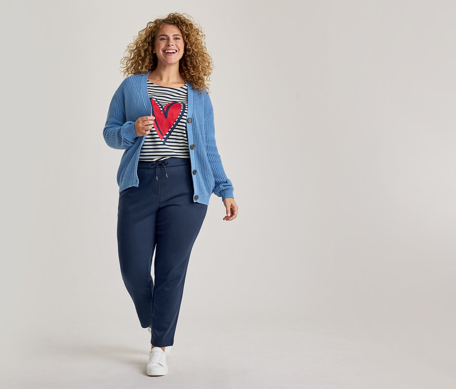 Une femme souriante aux cheveux bouclés marche vers l'avant, vêtue d'un cardigan bleu en maille, d'un t-shirt rayé avec un cœur rouge, d'un pantalon de jogging bleu marine et de baskets blanches.