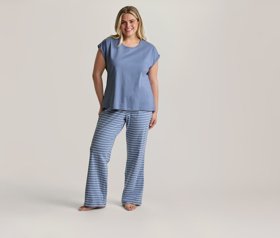 Femme blonde souriante debout pieds nus, portant un ensemble de pyjama composé d'un t-shirt bleu et d'un pantalon rayé bleu et blanc.