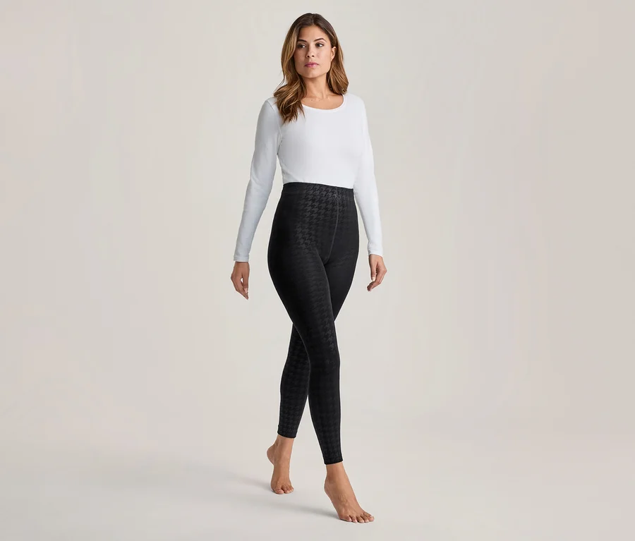 Femme portant un haut blanc à manches longues et des leggings noirs à motif pied-de-poule, debout pieds nus.