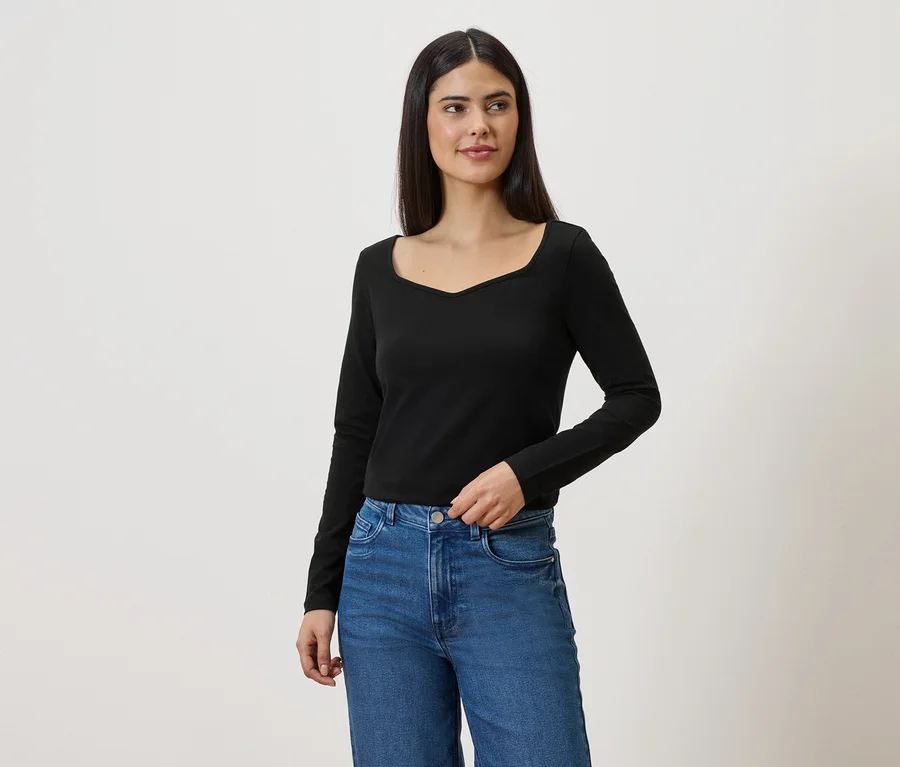 Une femme aux longs cheveux foncés porte un chemisier noir à manches longues et un jean bleu sur fond blanc.