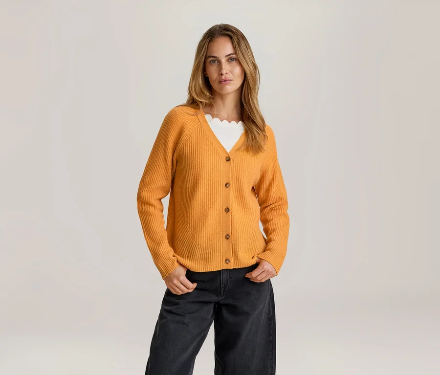 Femme aux longs cheveux châtains clairs, debout les mains dans les poches, portant un cardigan Mérinos orange ouvert sur un top blanc à bord festonné et un pantalon large foncé.