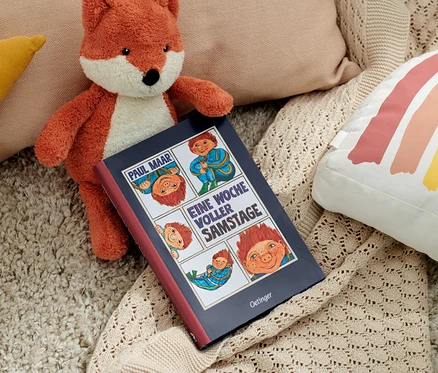 Un renard en peluche est posé à côté du livre « Das Sams – une semaine pleine de samedi » et de coussins.