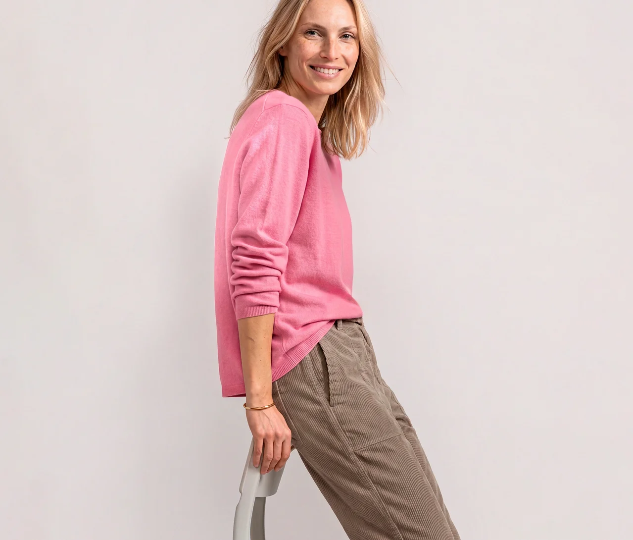 Femme souriante portant un pull en maille Street One rose pâle et un pantalon en velours côtelé beige, appuyée sur une chaise.