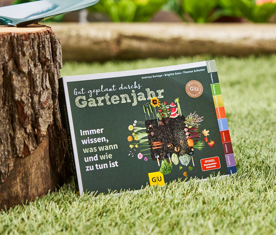 Le livre « Gut geplant durchs Gartenjahr » est posé sur l'herbe à côté d'une souche d'arbre sur laquelle repose une pelle de jardin.
