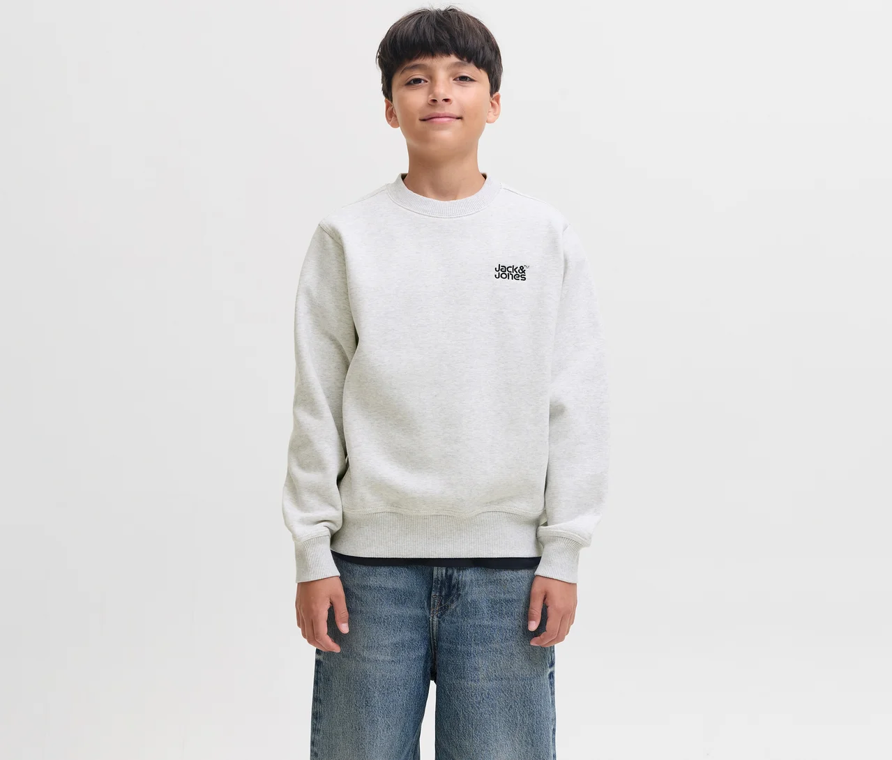 Un jeune garçon aux cheveux foncés sourit légèrement, portant un pull Colton blanc de Jack&Jones avec le logo de la marque sur la poitrine et un jean bleu.