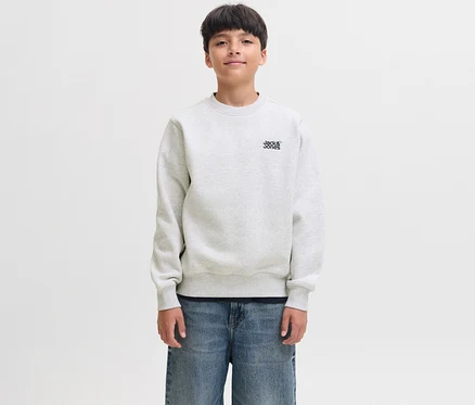 Un jeune garçon aux cheveux foncés sourit légèrement, portant un pull Colton blanc de Jack&Jones avec le logo de la marque sur la poitrine et un jean bleu.