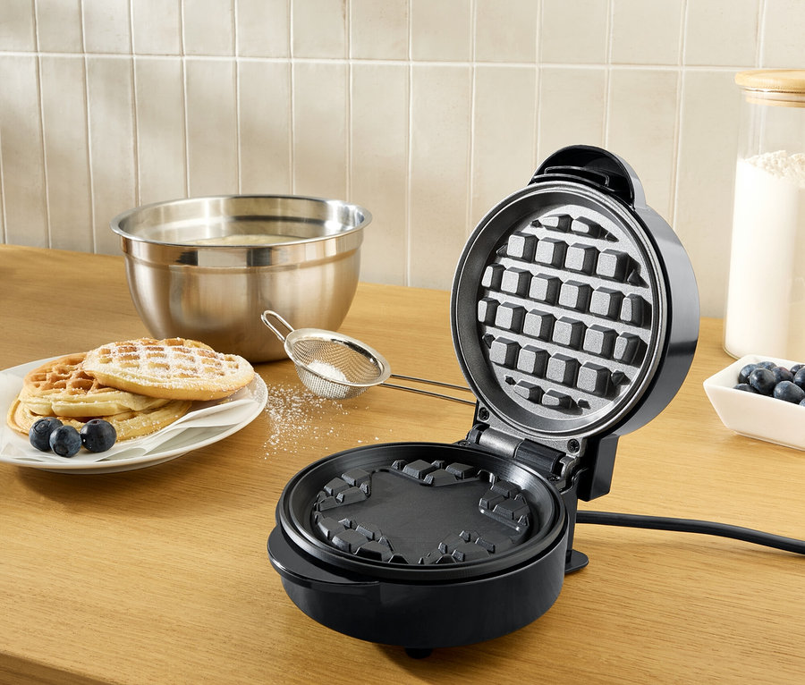 Un gaufrier noir ouvert sur un plan de travail en bois à côté d'une assiette de gaufres saupoudrées de sucre glace et de myrtilles.