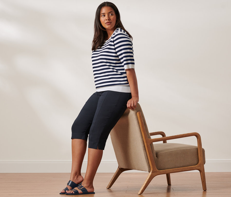 Une femme est assise sur le dossier d'une chaise et porte un pull en maille fine bleu et blanc rayé, un corsaire stretch et des sandales à brides.