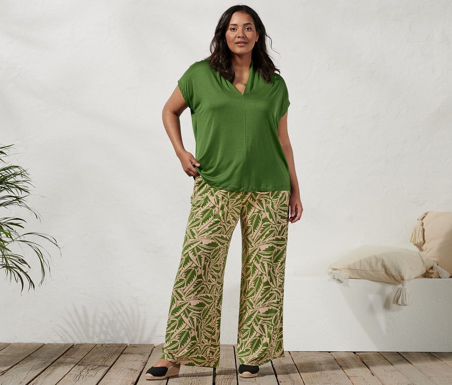 Une femme debout porte un t-shirt à col montant vert, un pantalon palazzo imprimé vert beige et des espadrilles à talon compensé.