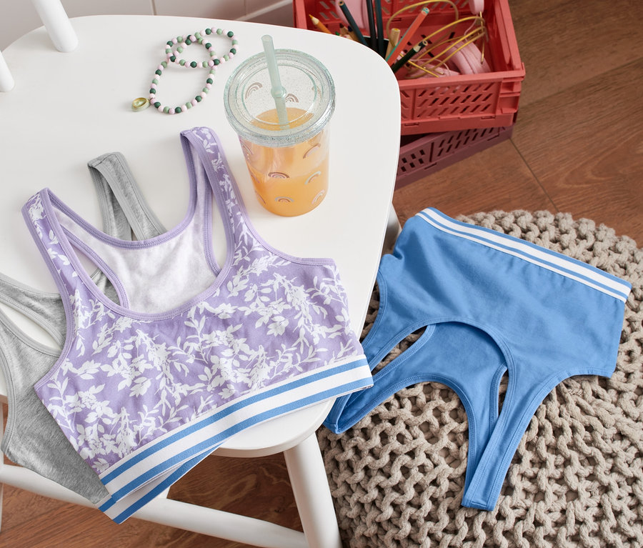 Trois brassières sont posées sur une chaise blanche : une grise, une violette à motif floral et une bleue. À côté d’elles se trouve un verre avec une boisson.