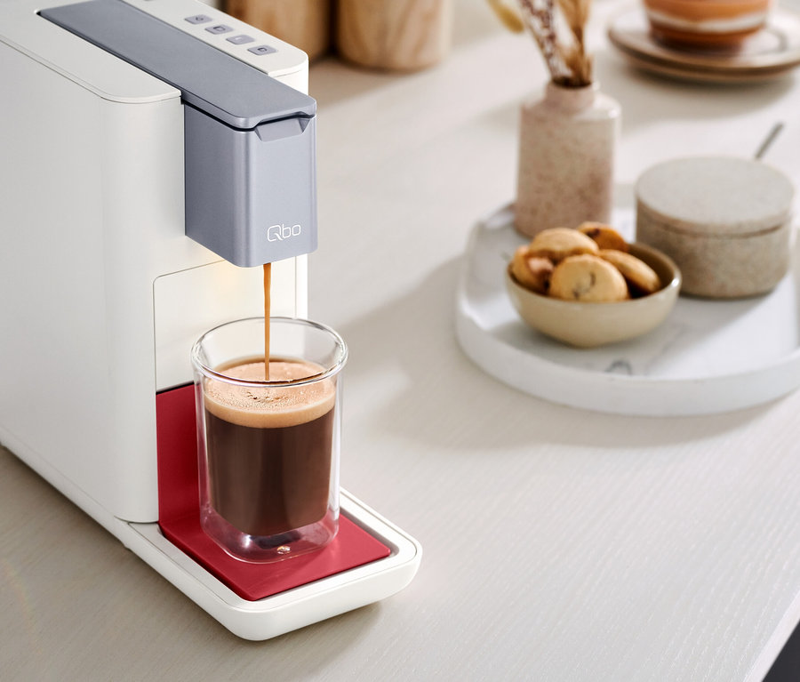 Le café coule d'une machine à café Qbo ESSENTIAL Bright Coral dans un verre.