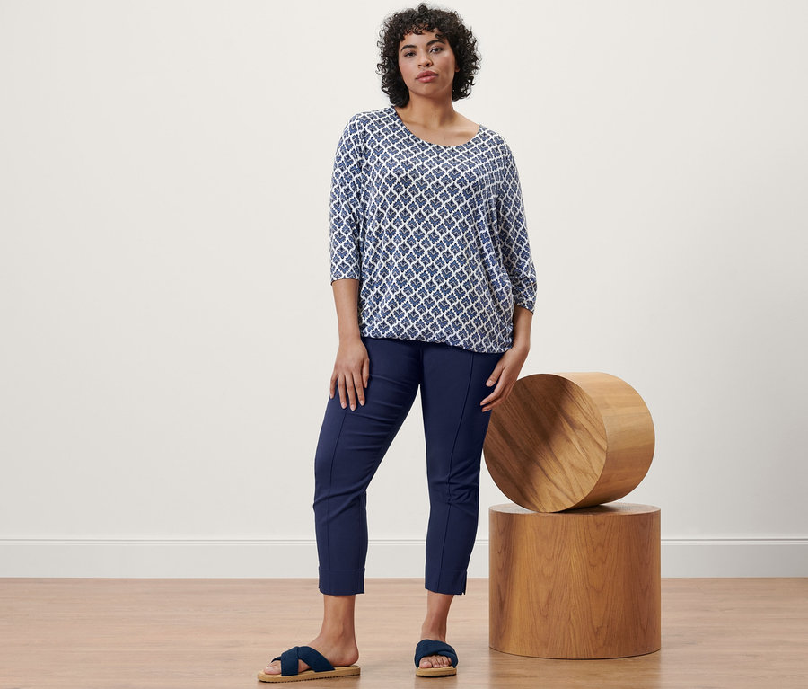 Une femme debout portant une blouse imprimée, un pantalon stretch bleu foncé et des sandales confortables.