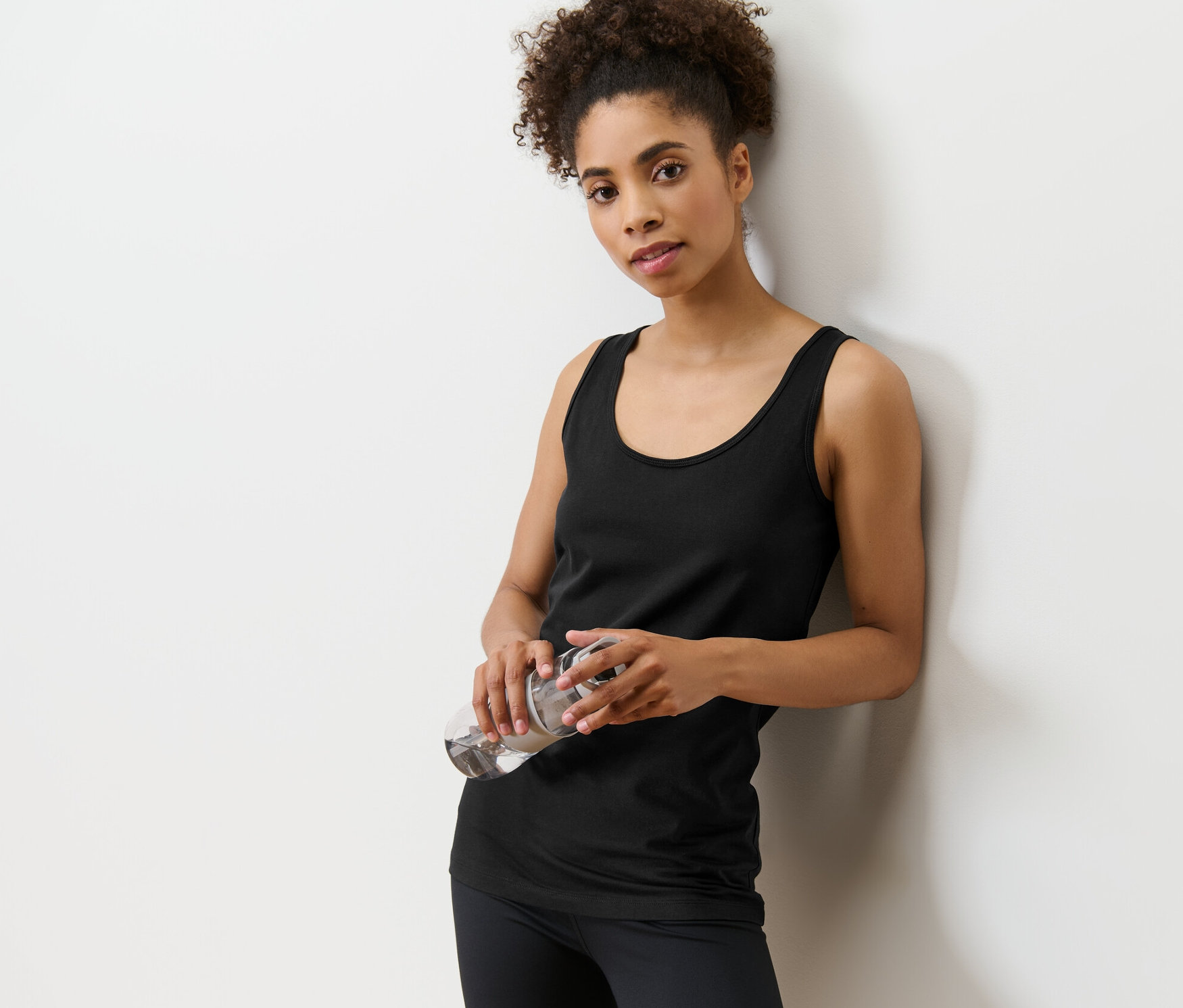 Femme appuyée contre un mur tenant une bouteille d'eau. Elle porte un débardeur noir d'un ensemble de 2.