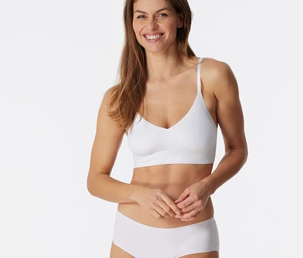 Femme souriante aux cheveux bruns portant une brassière invisible SCHIESSER blanche avec coussinets et un slip blanc, mains jointes.