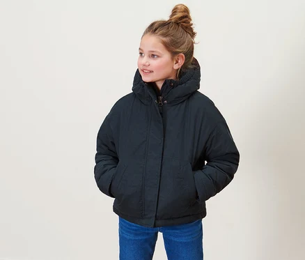 Une fille debout porte une veste de mi-saison pour enfants, un jean bleu et des chaussures marron.