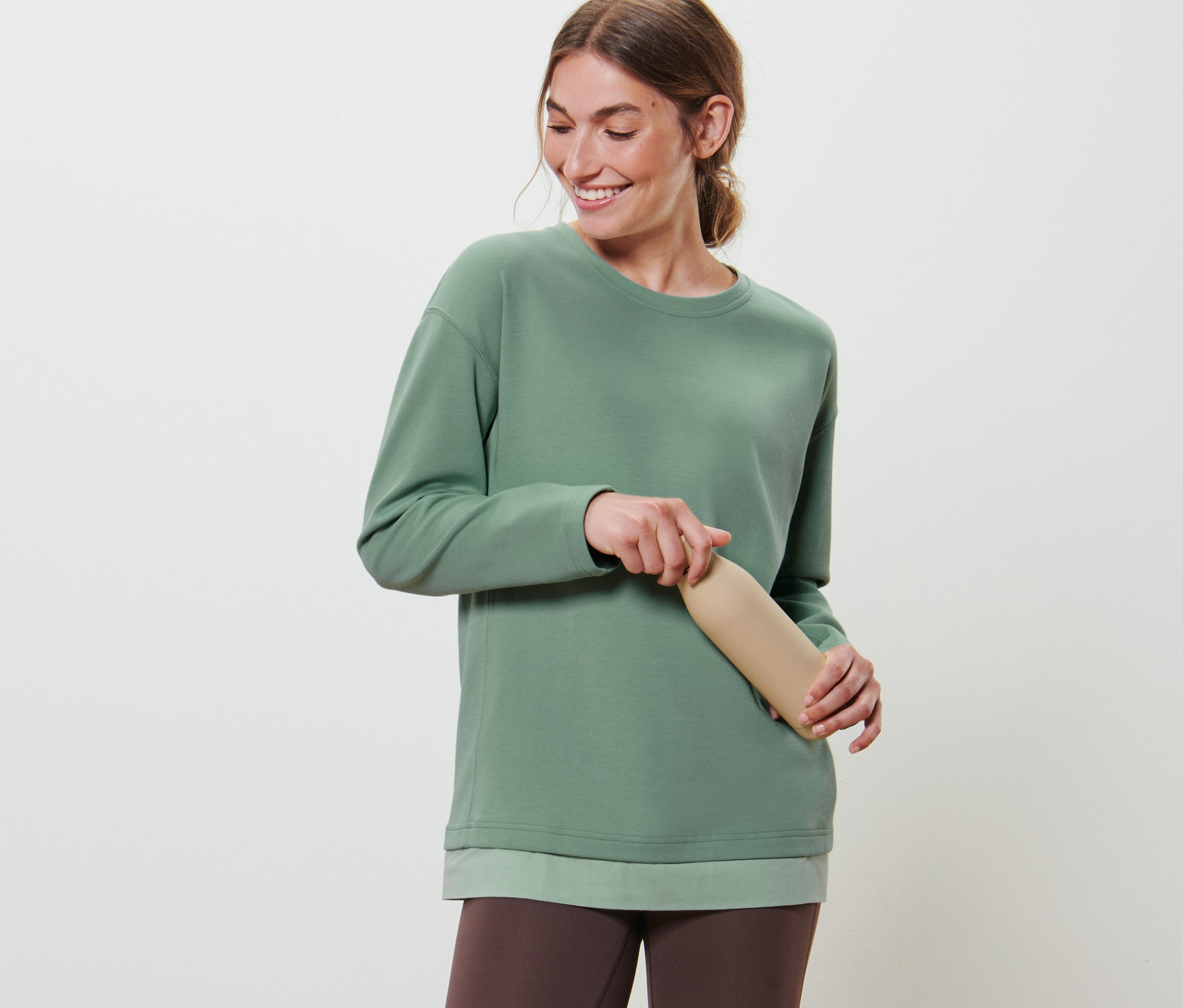 Femme tenant une bouteille d'eau beige et portant un haut à manches longues fitness oversize vert et un legging marron.