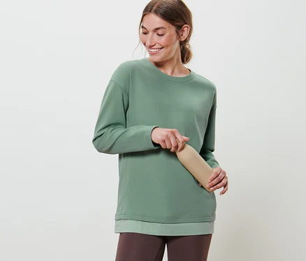 Femme tenant une bouteille d'eau beige et portant un haut à manches longues fitness oversize vert et un legging marron.