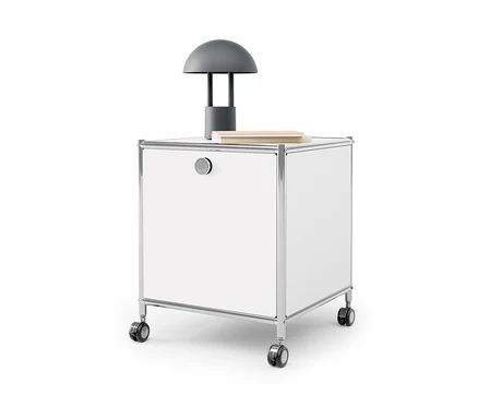 Table d'appoint blanche avec tiroir en métal « CN3 » avec une lampe grise et un livre dessus.