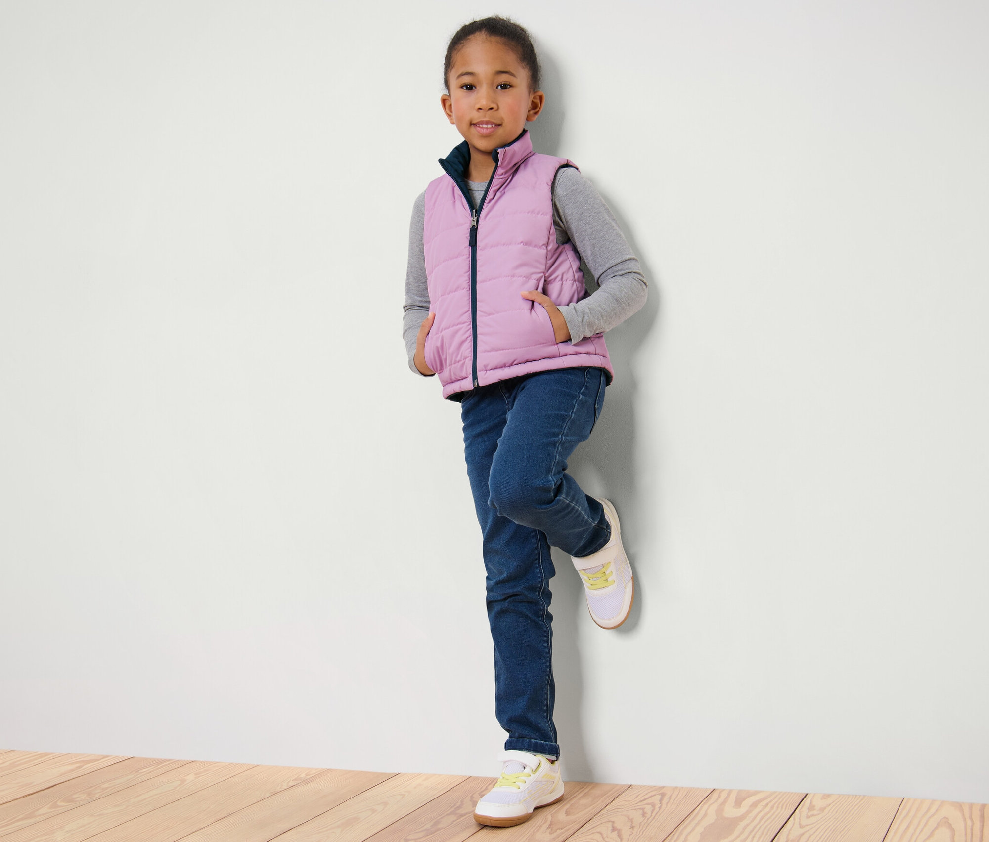 Une fille appuyée contre un mur porte un gilet sans manches matelassé réversible pour enfant, un jean et des baskets.