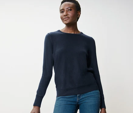 Une femme pose en pull en maille fine de mérinos bleu foncé et un jean à côté d'un tabouret en bois.