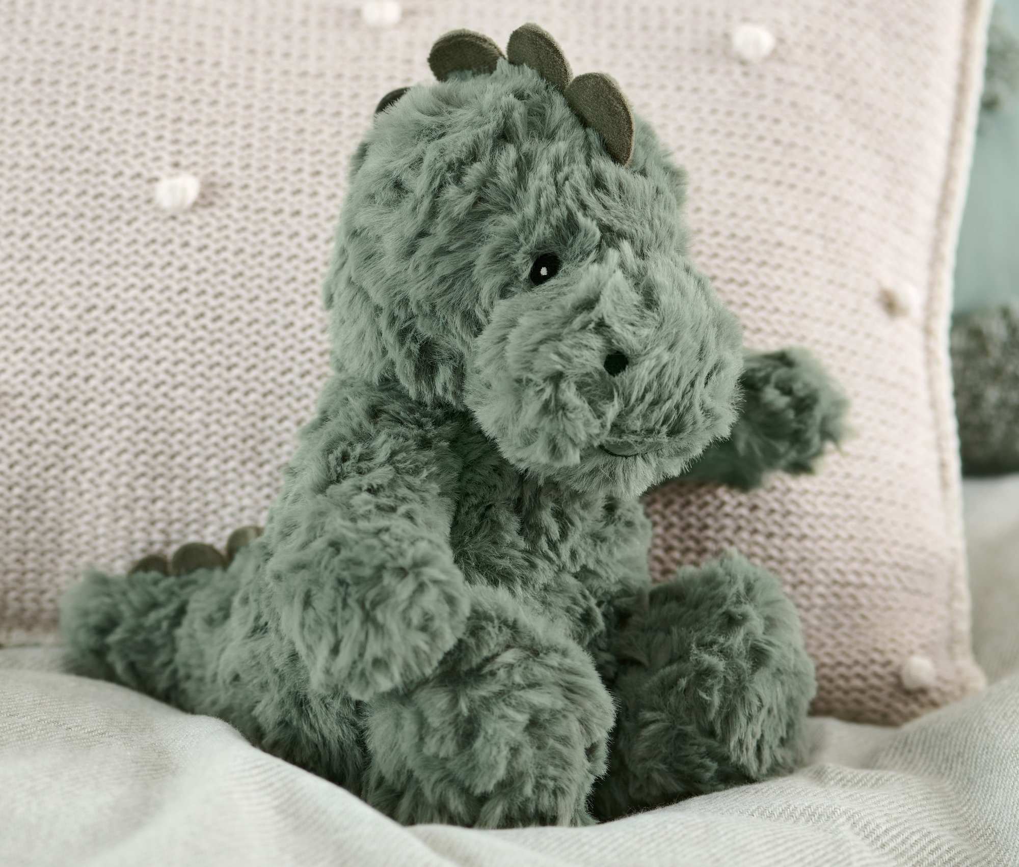 Un dinosaure en peluche est assis sur un lit à côté d'un oreiller beige.