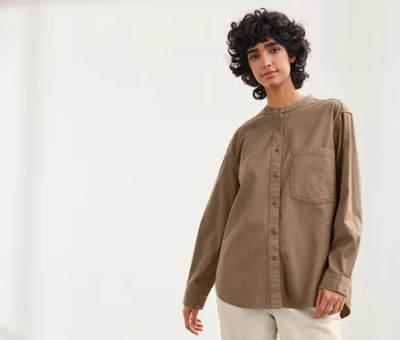 Une femme pose sur un fond clair, portant une blouse NAH/STUDIO en coton durable couleur noisette et un pantalon blanc.
