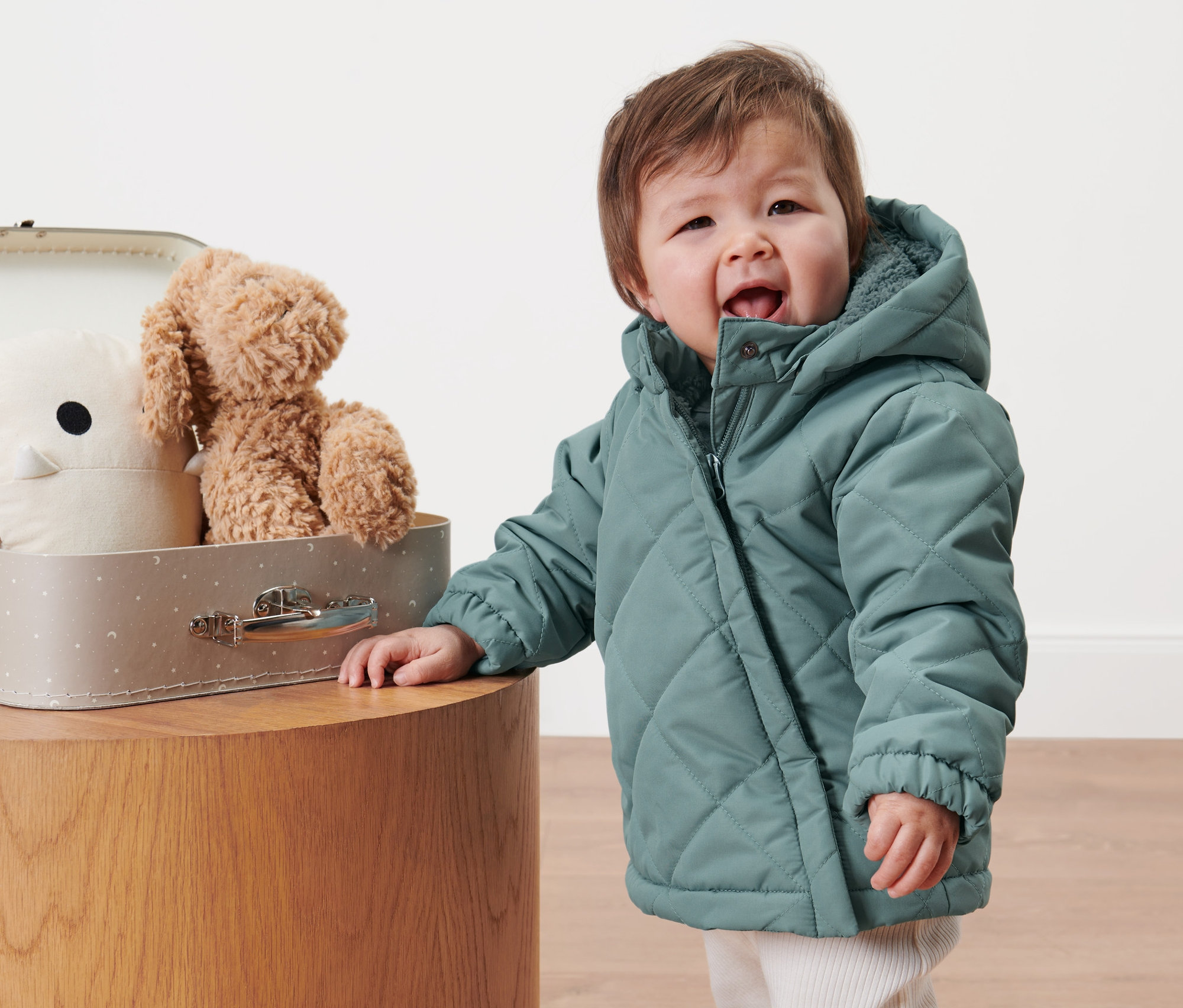 Un jeune enfant portant une veste matelassée pour tout-petit se tient à côté d'une valise contenant des jouets.