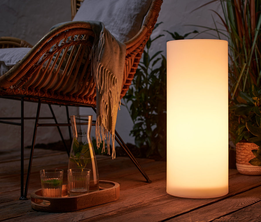 Sur une terrasse en bois se trouve une colonne lumineuse solaire avec changement de couleur, env. 50 cm, à côté d'un fauteuil en osier avec coussin.