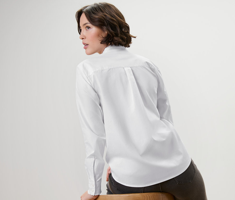 Une femme assise de côté sur une chaise en bois porte un chemisier blanc avec des détails ruchés.