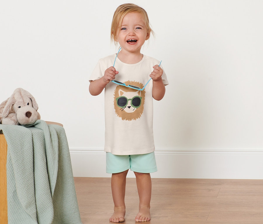 Un enfant souriant tient des lunettes de soleil et porte un t-shirt pour enfant avec un motif en paillettes réversibles. Un chien en peluche est posé sur un tabouret en bois recouvert d'une couverture.