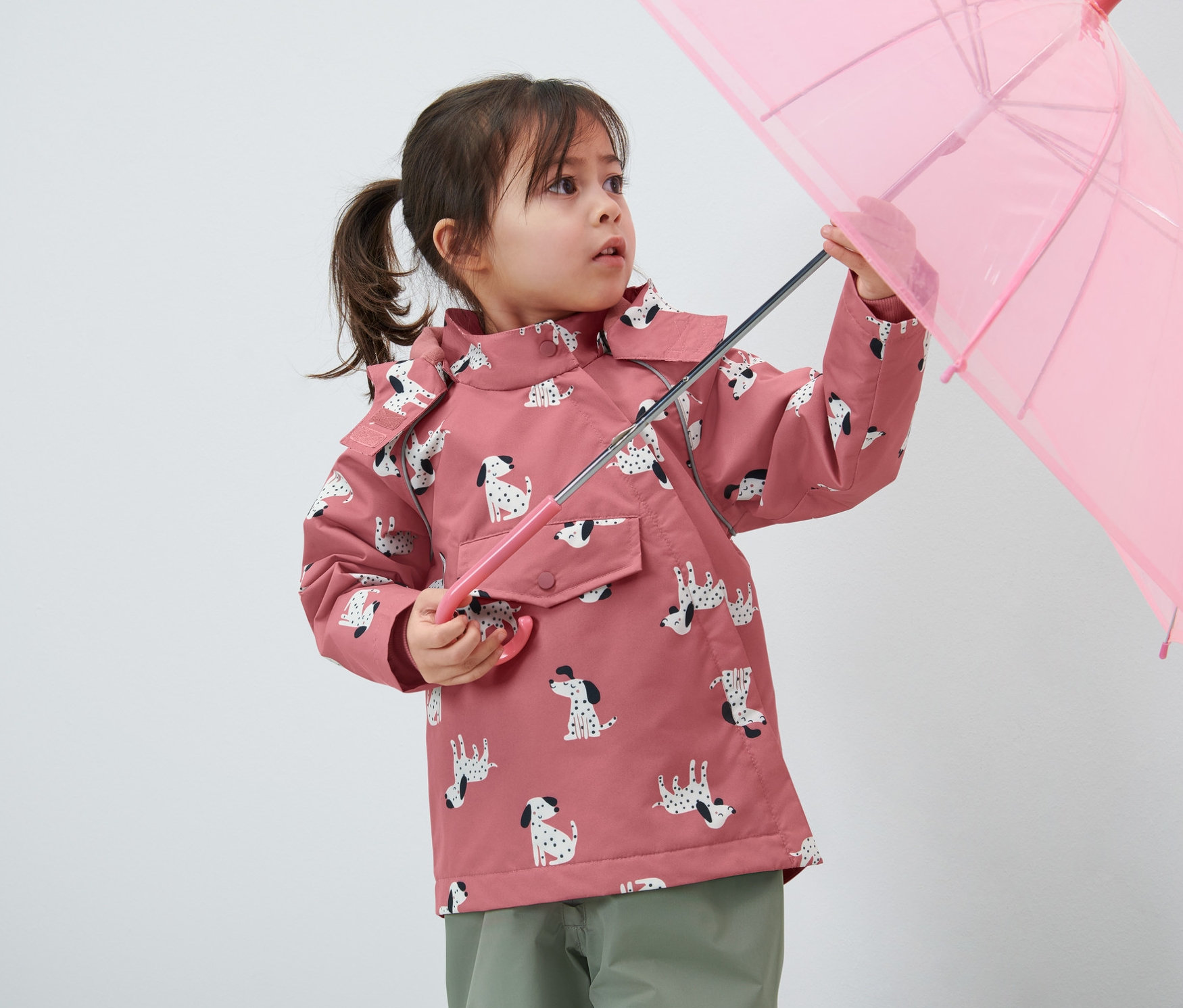 Une petite fille tient un parapluie rose et porte une veste de pluie thermique pour enfant avec un motif de chiens.