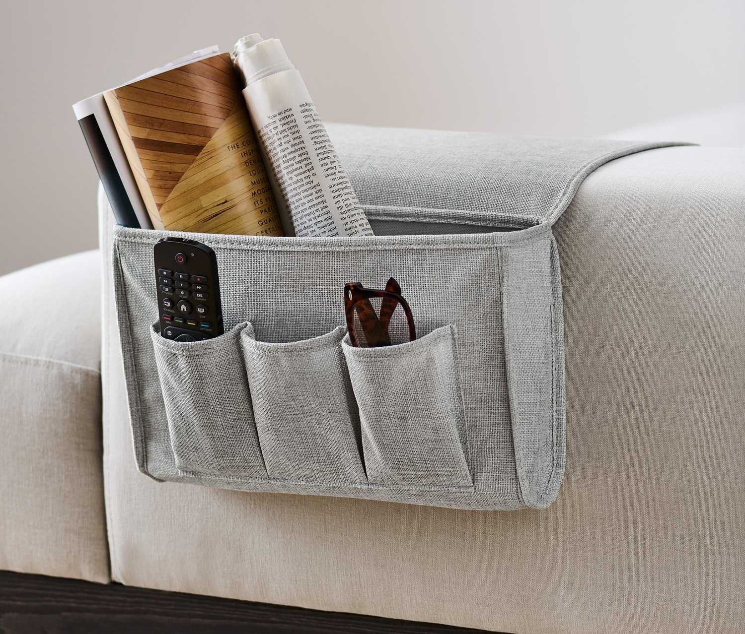 Un rangement de lit ou de canapé gris est accroché sur le côté d'un canapé et contient une télécommande, des lunettes, des livres et un journal.