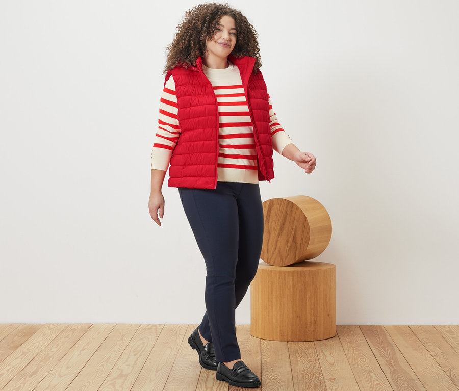 Une femme marche et porte un pull en maille fine rayé rouge, un gilet sans manches matelassé à capuche et un pantalon en bengaline bleu foncé.