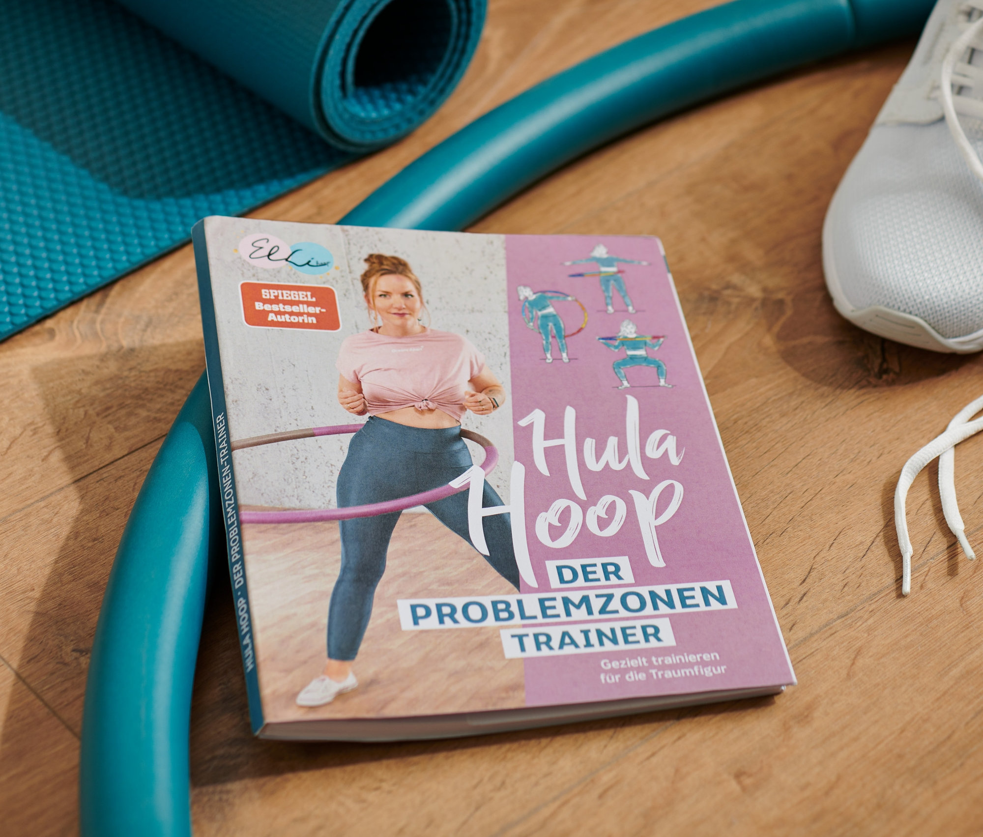 Sur un plancher en bois se trouvent un livre de hula hoop, un tapis d'entraînement bleu, un cerceau bleu et des baskets blanches.