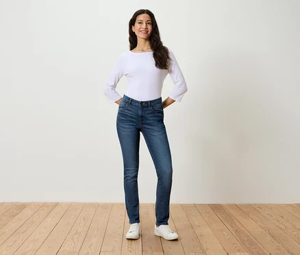 Une femme se tient debout, les mains sur les hanches, portant un jean bleu, un chemisier blanc et des baskets blanches.