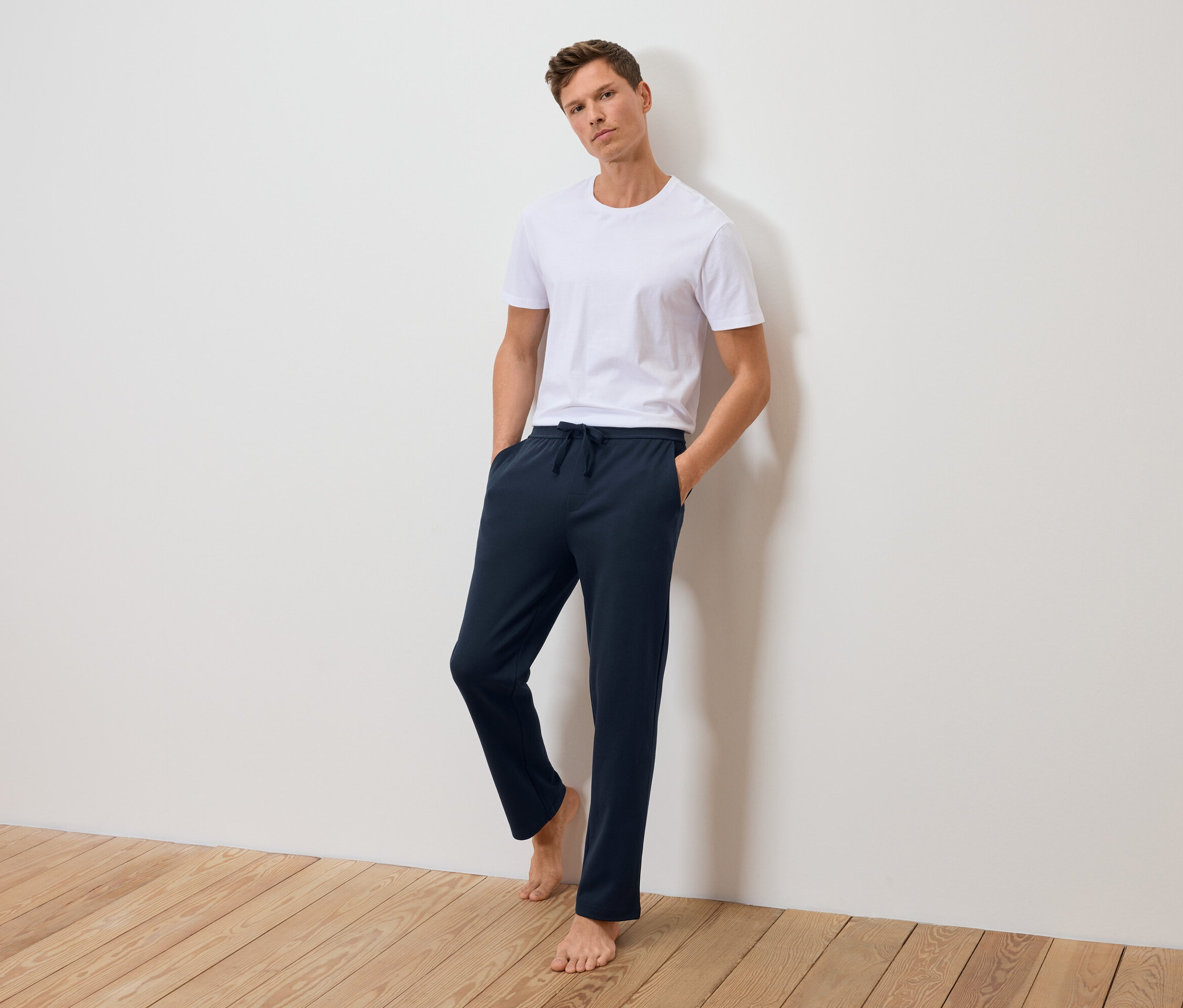 Un homme est appuyé contre un mur, portant un t-shirt blanc et un pantalon de survêtement bleu marine.