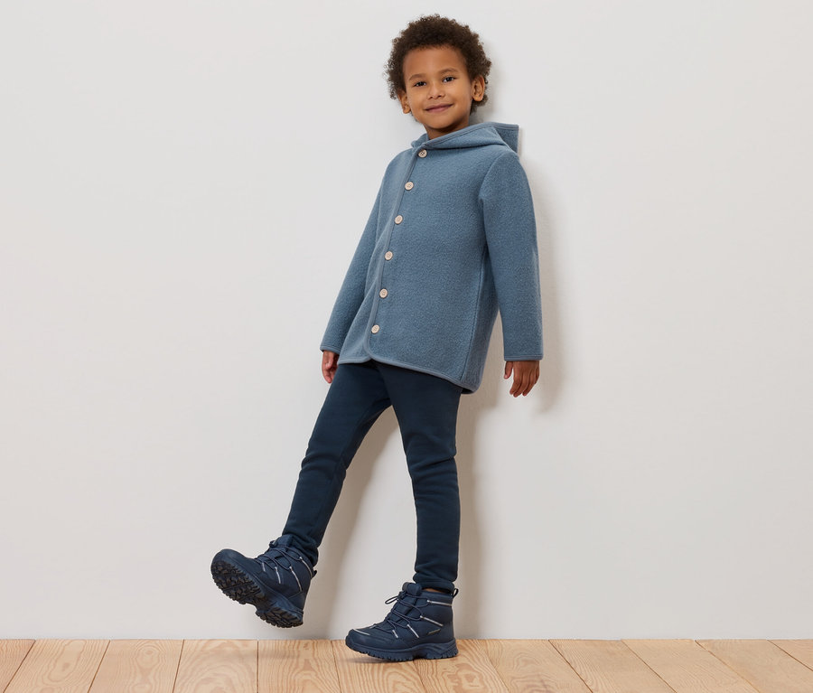 Un garçon posant contre un mur, portant un manteau en laine bleu à capuche, un jean bleu foncé et des bottes d'hiver bleues.