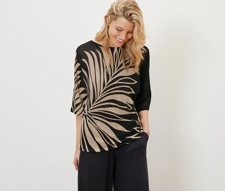 Une femme blonde sourit et a les mains dans les poches d'un pantalon noir ample. Elle porte un pull-over en maille fine imprimé.
