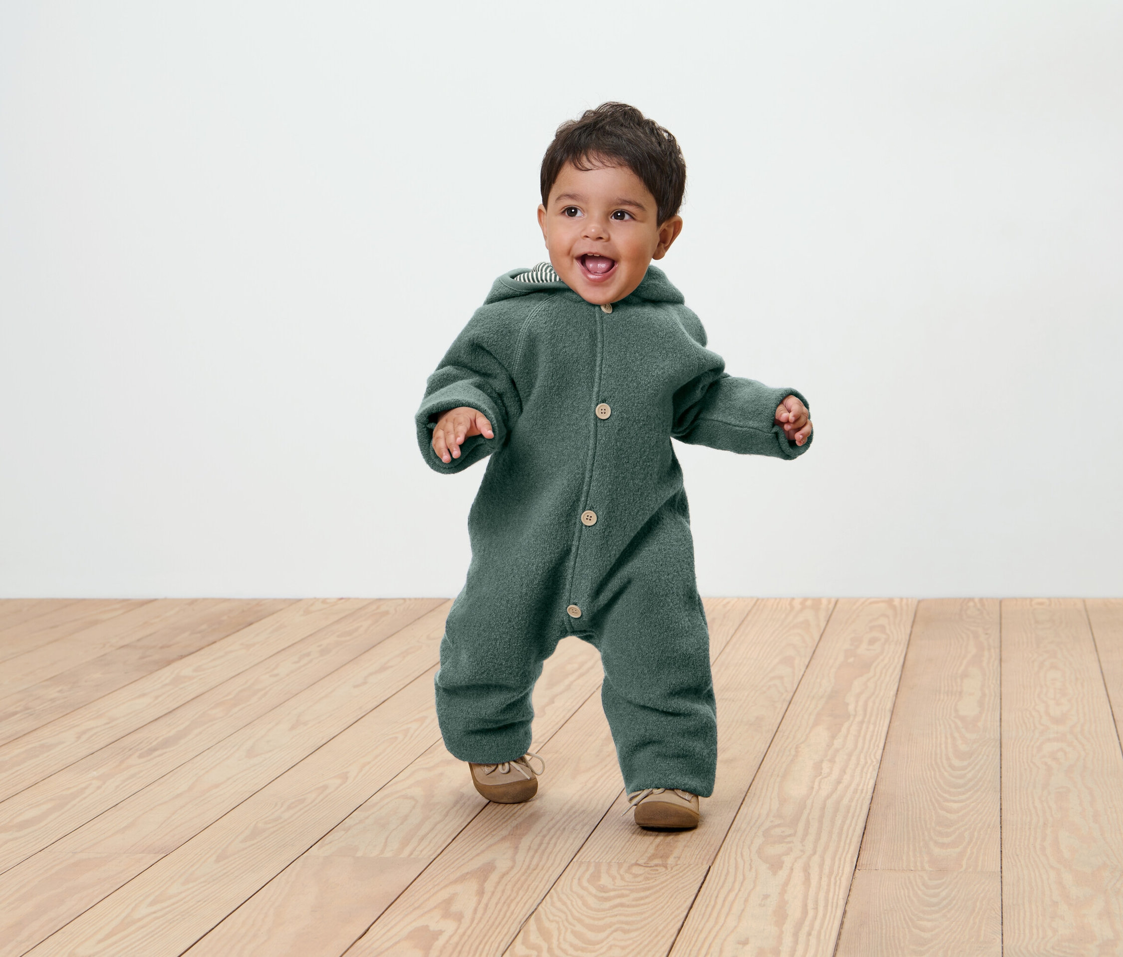 Un bébé souriant vêtu d'une combinaison verte à capuche et de chaussures sur un plancher en bois.