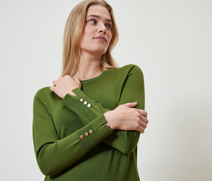 Blonde portant un pull olive avec des boutons dorés sur les manches, les bras croisés.