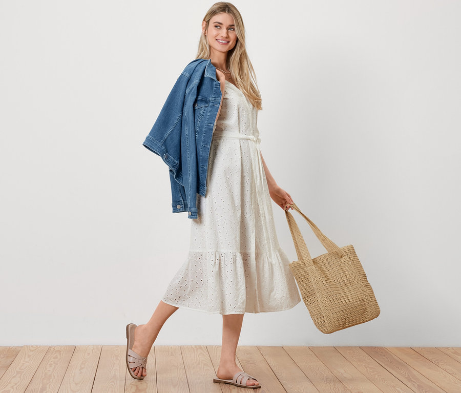 Une femme marche avec une veste en jean sur l'épaule, un cabas aspect raphia et une robe avec broderie ajourée.