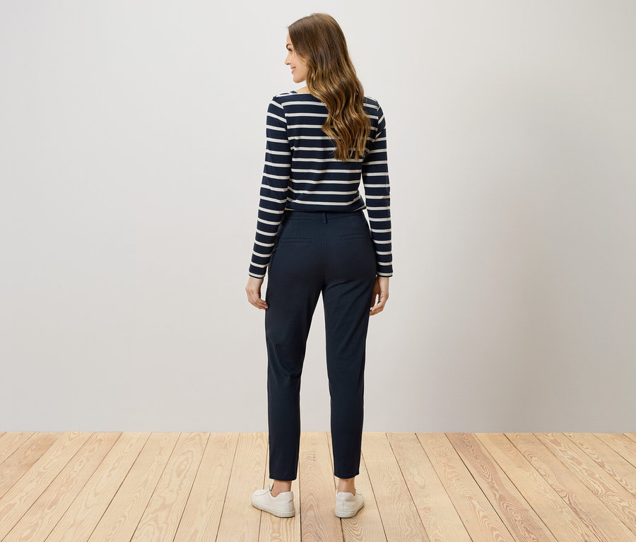 De dos, une femme aux cheveux bruns porte un pantalon bleu marine et un t-shirt rayé.