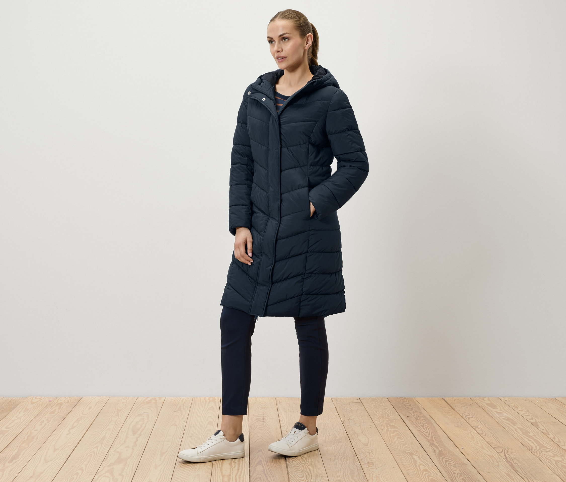 Une femme pose dans un long manteau matelassé bleu marine, un pantalon bleu marine et des baskets blanches.