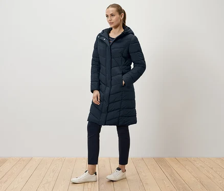 Une femme pose dans un long manteau matelassé bleu marine, un pantalon bleu marine et des baskets blanches.