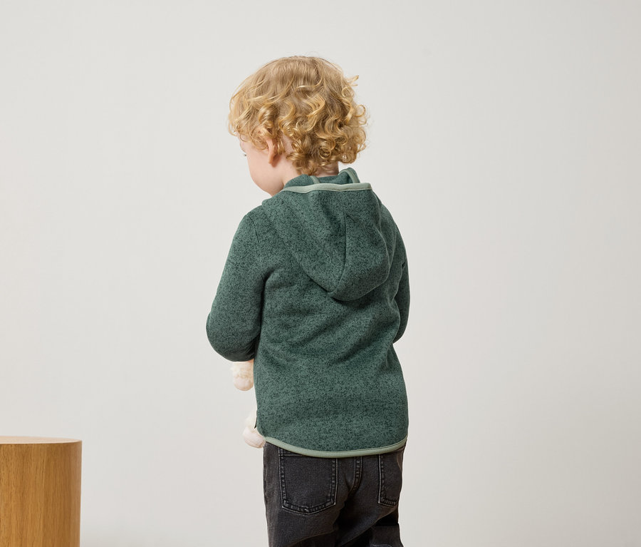 Un garçon aux cheveux blonds et bouclés se tient dos à la caméra, portant un sweat à capuche vert et un jean noir.