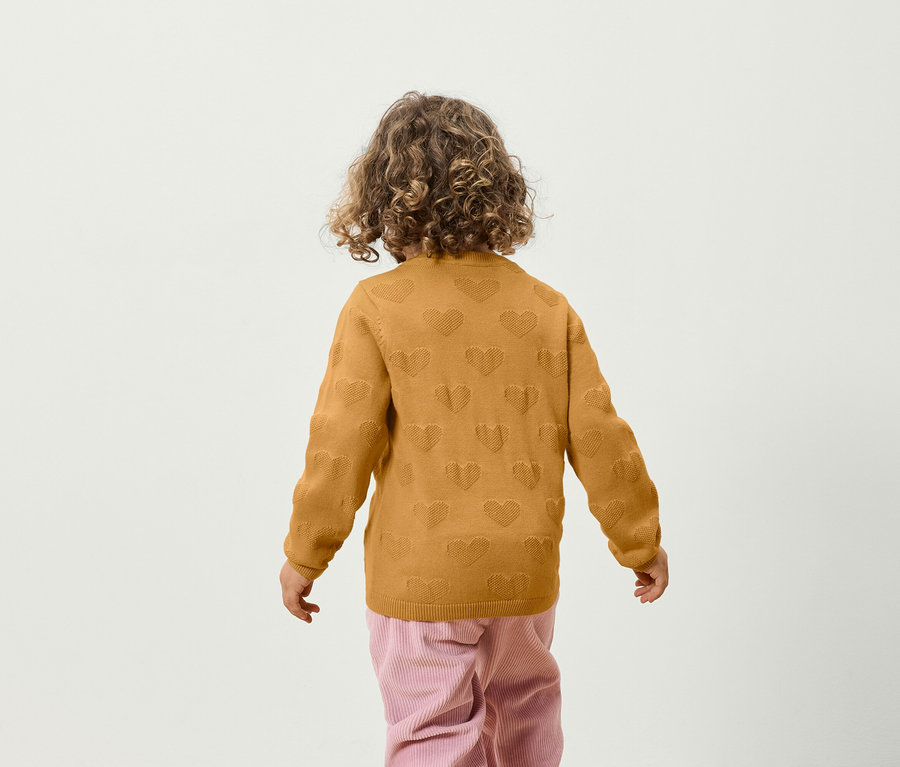 Un garçon aux cheveux bouclés se tient dos à la caméra, portant un pull jaune avec un motif de cœurs et un pantalon rose.