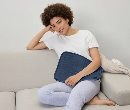 Une femme est assise sur un canapé et tient un coussin chauffant bleu sur ses genoux.