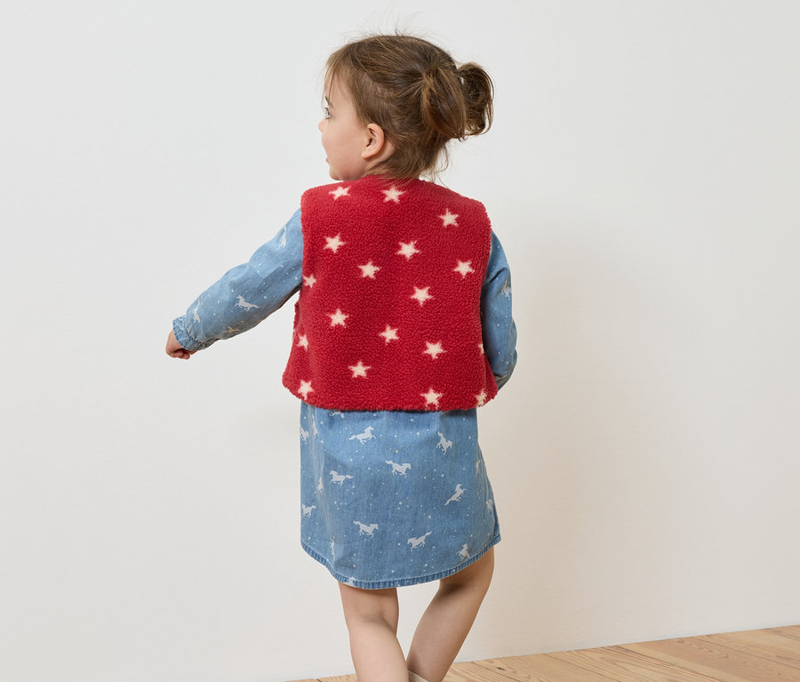 Une fillette portant une robe en jean avec des imprimés de chevaux et un gilet rouge à étoiles blanches, vue de dos.
